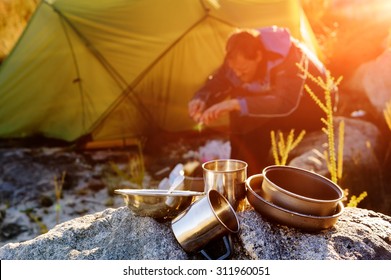 Adventure Camping Man Cooking Alone Outdoors With Tent, Sunrise And Lens Flare In The Mountain Morning Sunlight. Happy Explorer Enjoying A Meal In The Wilderness