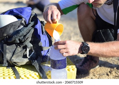 Adventure Man Pouring Hydration Electrolyte Powder In Water Bottle. A Backpack And A Bottle Of Water With Powdered Electrolyte On Desert Mountains Ground. Man Traveler Preparing Isotonic Drink 