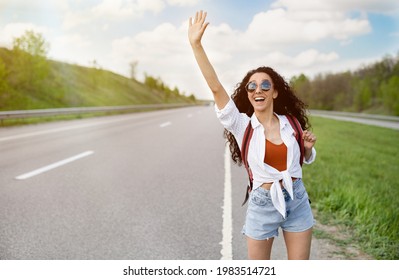 Adventure and tourism concept. Beautiful millennial woman in casual wear waving down car, hitchhiking on roadside, copy space. Pretty young lady traveling by autostop, having summer trip