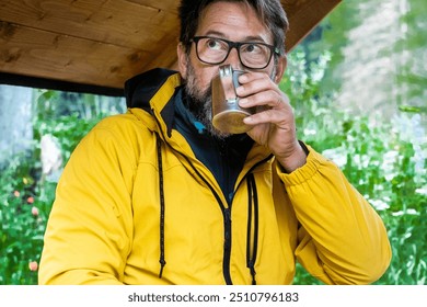 Adventure travel vacation people lifestyle. One adult man drinking cup of coffee sitting on a wooden table bench in park mountain scenic destination. Mature man enjoying journey. Wearing yellow jacket - Powered by Shutterstock