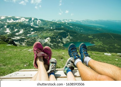 Adventures In The Rila Mountains Of Bulgaria. Family On Vacation. Tourists Enjoy The View Of The Mountains And The Lake. Top View On Legs