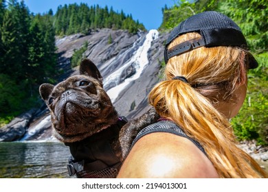 Adventurous athletic female hiker standing at the base of a waterfall with her French Bulldog in a backpack in the Pacific Northwest.