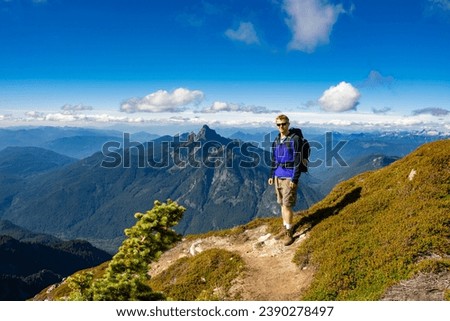 Adventurous athletic male hiker standing on a hiking trail on top of a rugged mountain at at the camera smiling, in the Pacific Northwest with jagged mountains in the background.

