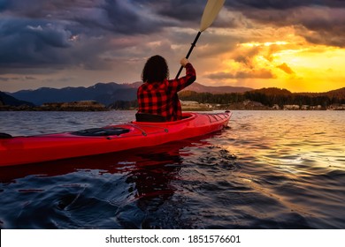 Adventurous Girl Paddling On A Bright Red Kayak In Calm Ocean Water. Dramatic Colorful Sunrise Sky. Taken In Indian Arm, Deep Cove, North Vancouver, British Columbia, Canada.