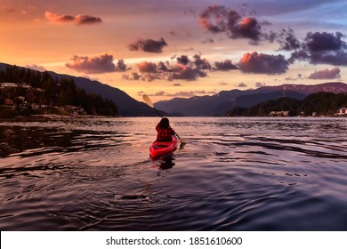 Adventurous Girl Paddling On A Bright Red Kayak In Calm Ocean Water. Dramatic Colorful Sunset Sky. Taken In Indian Arm, Deep Cove, North Vancouver, British Columbia, Canada.