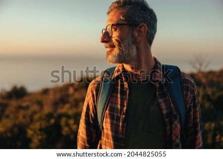 Adventurous male hiker looking away while standing on a hilltop. Happy mature hiker standing with a backpack on outdoors. Cheerful mature man enjoying the sunset and view.