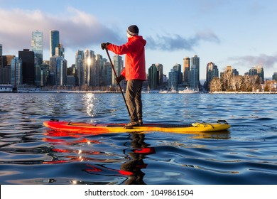 Adventurous Man Is Paddle Boarding Near Downtown City During A Vibrant Winter Sunrise. Taken In Coal Harbour, Vancouver, British Columbia, Canada. Concept: Adventure, Travel, Holiday, Lifestyle