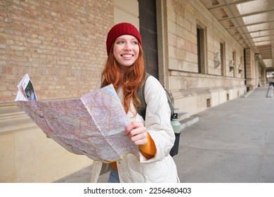 Adventurous redhead girl walks in town with paper map, explores city as tourist, looks for popular tourism attractions, looks around excited and smiles.