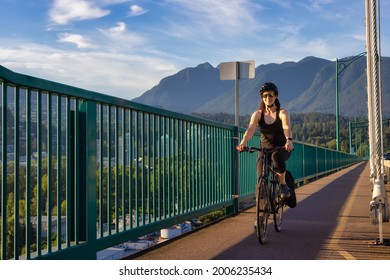 Adventurous White Caucasian Adult Woman riding a road bicycle on a bike path across the famous Lions Gate Bridge in a modern city. Sunny Summer. Downtown Vancouver, British Columbia, Canada.
