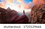 Adventurous Woman at the edge of a cliff is looking at a beautiful landscape view in the Canyon during a vibrant sunset. Taken in Zion National Park, Utah, United States. Sky Composite Panorama