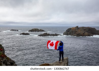 Adventurous woman holding a Canadian Flag on a Rocky Atlantic Ocean Coast during a cloudy day. Taken in Sleepy Cove, Crow Head, Twillingate, Newfoundland, Canada.