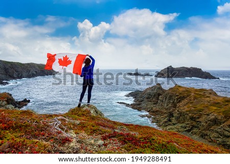 Adventurous woman holding a Canadian Flag on a Rocky Atlantic Ocean Coast during a cloudy and blue sky day. Taken in Sleepy Cove, Crow Head, Twillingate, Newfoundland, Canada.