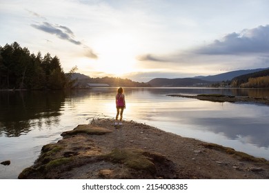 Adventurous Woman on a shore enjoying colorful Sunset. Shoreline Trail, Port Moody, Greater Vancouver, British Columbia, Canada. Park in Modern City.