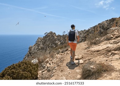 Adventurous Young Man Hiking Scenic Coastal Trail Along Rugged Cliffs Overlooking Azure Sea Under Clear Blue Sky - Powered by Shutterstock