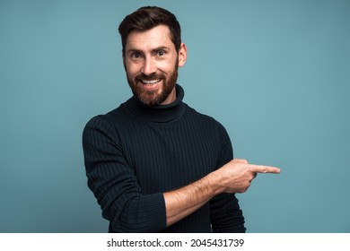 Advertise Here. Positive Bearded Man Pointing Finger Away Paying Your Attention At Empty Space For Advertisement, Looking At Camera With Toothy Smile. Indoor Studio Shot Isolated On Blue Background 