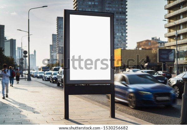 Advertising billboard for commercial images and\
videos standing on sidewalk near road. Drivers in cars, passengers\
in public transport and pedestrians bassing by banner. Busy\
traffic, houses,\
subway.