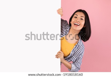 Advertising Billboard. Portrait of excited asian woman hiding behind blank white poster with mockup template and copy space for logo, design or message. Lady peeking out, pink studio wall