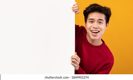 Advertising Billboard. Portrait Of Smiling Asian Guy Hiding Behind Blank White Poster With Mockup Template And Copy Space For Logo, Design Or Message. Male Teen Peeking Out, Orange Studio Wall