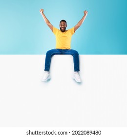 Advertising Billboard Template. Young Black Guy Sitting On Top Of Blank White Poster With Mockup, Lifting Hands Up In Excitement On Blue Studio Background, Space For Design