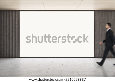 Advertising concept with businessman walking by blank white glowing screen with space for your logo or text in empty hall with light wooden ceiling and grey concrete floor, mockup