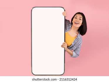 Advertising Concept. Portrait Of Excited Asian Lady Peeking Out Big Giant Vertical Smartphone With White Blank Screen Presenting Device Display. Gadget With Empty Free Space For Mock Up, Pink Wall