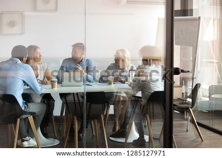 Advertising department brainstorming at modern office boardroom behind closed doors, view through the glass wall. Diverse staff led by ceo discussing new project sharing ideas thoughts and sales pitch