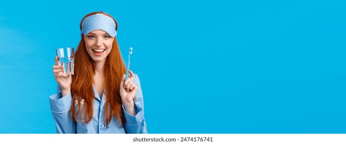 Advertising, hygiene and people concept. Feminine cute redhead girl feeling upbeat and enthusiastic start day right, drinking water, holding glass and toothbrush, wear nightwear and sleep mask. - Powered by Shutterstock