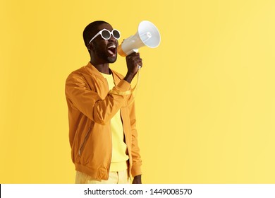 Advertising. Man screaming announcement in megaphone on yellow background. Portrait of african american male model in fashion wear using loud speaker in studio