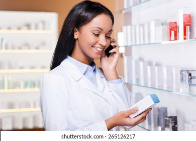 Advising A Proper Medicine. Beautiful Young African Woman In Lab Coat Holding Container With Some Medicine And Talking On The Mobile Phone While Standing In Drugstore 