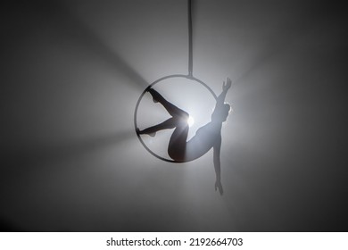 Aerial acrobat in the air ring. Young woman performs the acrobatic elements in the air hoop. Aerialist in on black background dark studio with backlight. For sports, acrobatic, circus school.