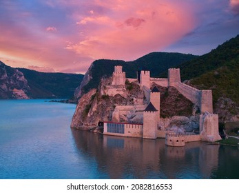 Aerial, Across Lake View Of The Medieval Fortress Golubac Over  Danube River. Fortress Towers Illuminated By Pink Light. Sunset, Pink And Red Clouds Sky. Outdoor And Traveling Theme. Serbia.