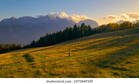 AERIAL: Athletic young lady is jogging along picturesque alpine meadow at sunset. Waning golden rays of autumn sun spill over scenic mountain landscape when she is running back down towards valley. - Powered by Shutterstock