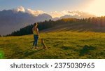 AERIAL: Attractive woman admires view of the mountains on a hilltop with her dog. Amazing sunny autumn day for outdoor recreation in the embrace of picturesque mountains and bonding time with pet.