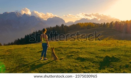 AERIAL: Attractive woman admires view of the mountains on a hilltop with her dog. Amazing sunny autumn day for outdoor recreation in the embrace of picturesque mountains and bonding time with pet.