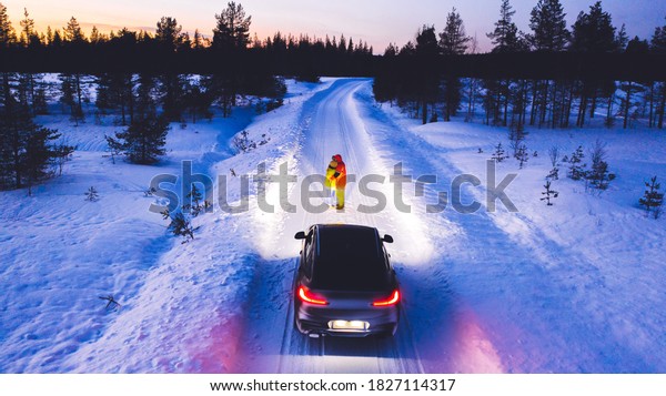 Aerial
back view of romantic couple in love of tourists standing near car
while headlights lighting them way on in winter darkness, vehicle
in snowy north lands. Wanderlust in road
trip
