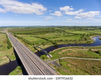 Aerial Birds Eye View Of A Busy Motorway With Bridge Crossing A River Of Water And Railway Track. Ennis County Clare Ireland.