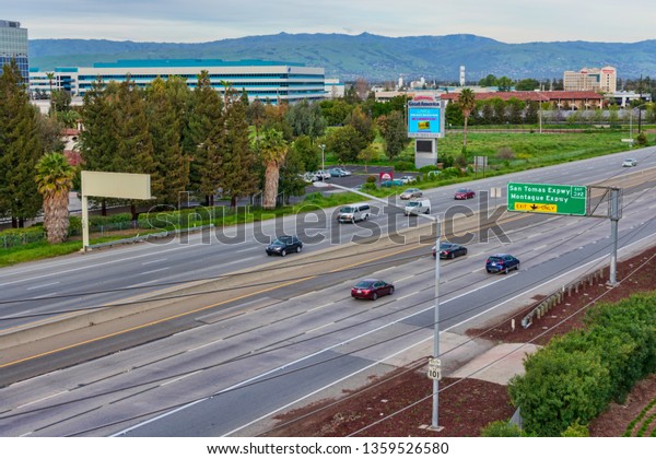Aerial bird\'s eye view of light weekend car traffic\
on eight-lane highway 101 in urban heart of Silicon Valley with\
green mountain range in background  - Santa Clara, California, USA\
- March 31, 2019