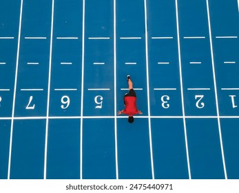 aerial bird's-eye view captured by a drone, this image depicts a runner crouched at the start line on a blue athletic track, poised to begin his training session. - Powered by Shutterstock