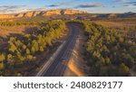 AERIAL: Black SUV car driving along the empty road through pine forest valley towards mesa tabletop mountains. Jeep on a journey across United States traveling through Utah landscape on sunny evening