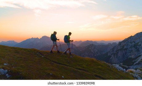 AERIAL: Carefree Active Woman And Man Enjoy A Calm Hike In The Serene Mountains Of Slovenia. Young Couple Hiking In The Beautiful Alps At Sunrise. Sporty Young Tourists Descend Down A Grassy Hill.