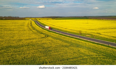 AERIAL: Cargo trucks driving on motorway road passing by yellow blooming fields. Running traffic on beautiful countryside asphalt roadway surrounded with flowering brassica rapa agricultural fields.