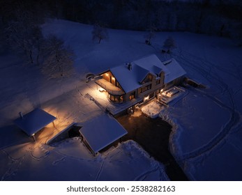 AERIAL: Cozy house covered with fresh snow glows warmly in quiet winter night. Inviting lights stand out against the frozen landscape, as snowy fields and trees surround the inviting country home. - Powered by Shutterstock