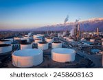 Aerial drone flight above an oil refinery at sunset with snow-covered a city and mountains in the background