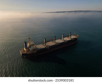 Aerial drone footage of a cargo ship sailing on misty waters with sunlight reflections, creating a calm and mysterious maritime scene, perfect for industrial and nautical themes - Powered by Shutterstock