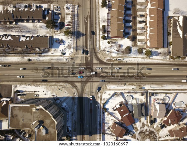 Aerial drone image of an intersection (junction)\
between two four lane roads in a north american suburb on a sunny\
day in winter.  Snow is on the ground and cars tracks can bee seen\
on the road.