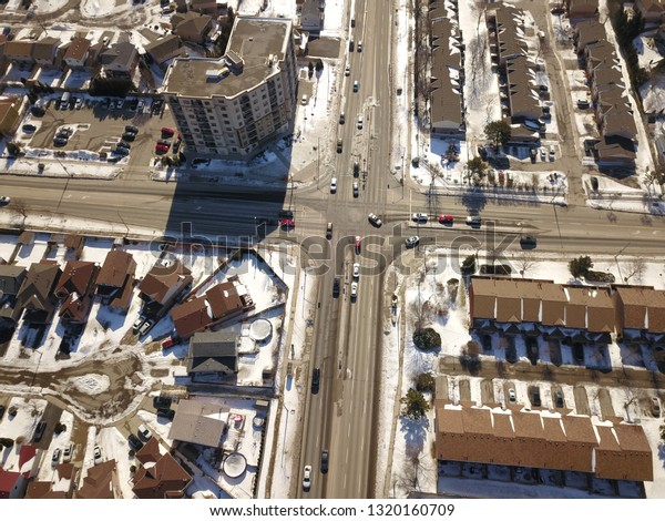 Aerial drone image of an intersection (junction)\
between two four lane roads in a north american suburb on a sunny\
day in winter.  Snow is on the ground and cars tracks can bee seen\
on the road.