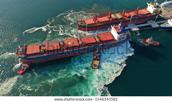 Aerial drone\
photo of tug boats manoeuvring industrial bulk carrier by pushing\
or pulling in Mediterranean\
refinery