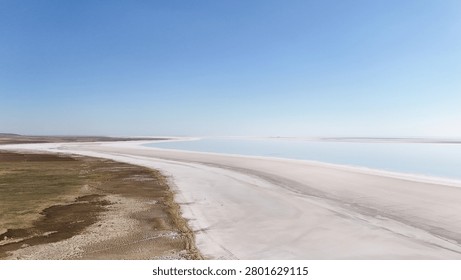 Aerial drone shot capturing the dramatic meeting point of two distinct ecosystems. On the right, a vast expanse of brilliant white salt flats stretches towards the horizon under a clear blue sky. On the left, a rugged shoreline of green vegetation and brown wetlands provides a stark textural and color contrast. The image highlights the natural division between the arid salt pan and the fertile marshland, showcasing the unique geology of the area.