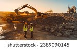 Aerial Drone Shot Of Construction Site On Sunny Evening: Caucasian Male Civil Engineer Talking To Hispanic Female Urban Planner and Using Tablet. Industrial Excavator Loading Rocks Into A Truck.