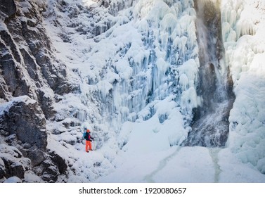 aerial drone shot of hiker with backpack, orange hat and orange pants standing next to waterfall surrounded by icicles and snow in winter - Powered by Shutterstock